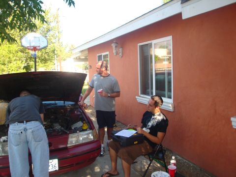 1993 Ford Taurus - Three men are gathered around the open hood of a red sedan, appearing to work on the engine.