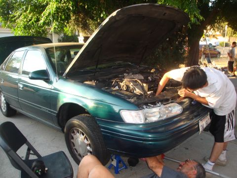 1993 Ford Taurus - Two people are working on the engine of a 1993 Ford Taurus, which is supported by jack stands.