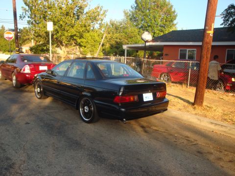 1993 Ford Taurus - A black sedan is parked on the street, with another red car visible in the background.