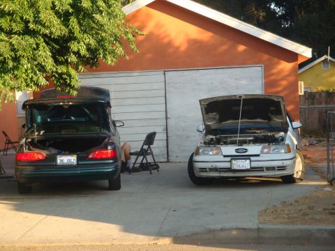 1993 Ford Taurus - A white 1993 Ford Taurus with its hood open is parked next to a dark sedan with its trunk open, where a person is working.
