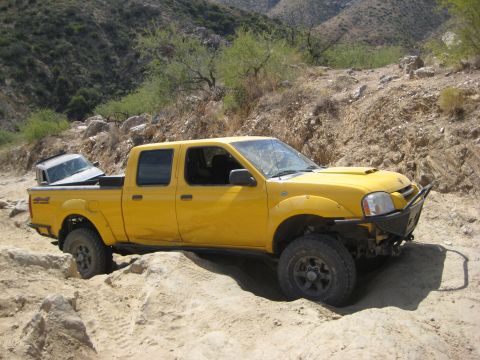 1996 Ford Bronco - A yellow pickup truck navigates a rocky, off-road trail with another vehicle loaded in its bed.