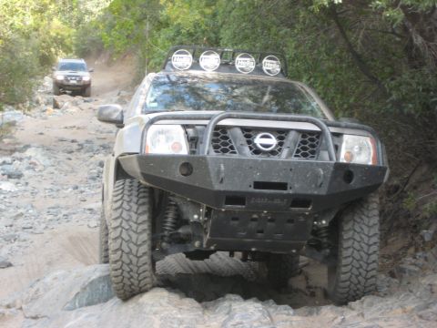 1996 Ford Bronco - Two SUVs navigate a challenging, rocky off-road path surrounded by trees.