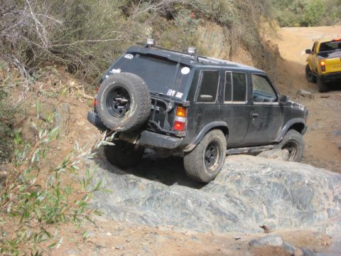 1996 Ford Bronco - A black SUV is seen off-roading on a rocky trail, with a yellow truck following in the background.