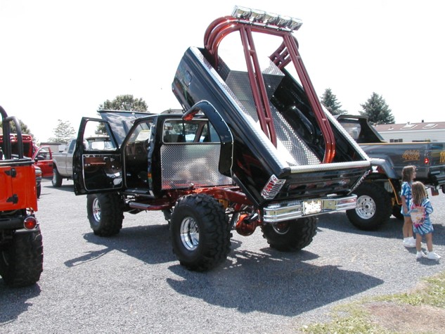 Modified pickup truck with dump bed raised, showing custom roll cage and diamond plate interior. Another truck and two girls are visible