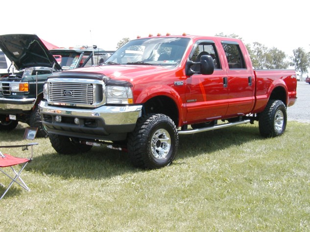 red Ford F-250 pickup truck with lifted suspension and chrome wheels, parked on grass next to another vehicle with hood open