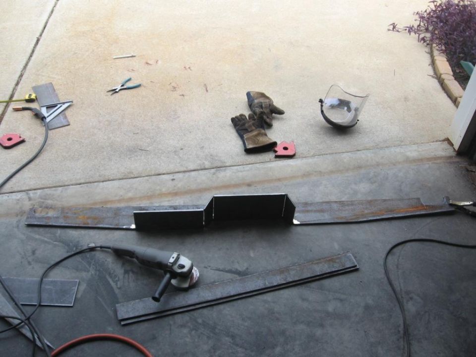 Metal pieces laid out on concrete with grinder, gloves, and face shield nearby. Looks like someone is building something custom