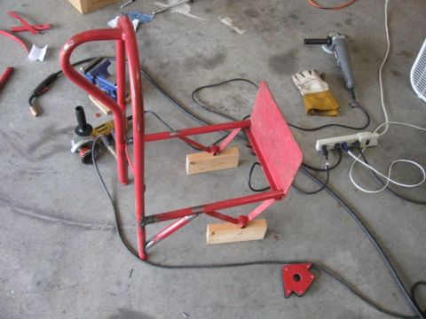 1995 Ford F-150 - A red metal frame, likely for a welding cart, rests on wooden blocks amidst various tools and electrical cords on a concrete floor.