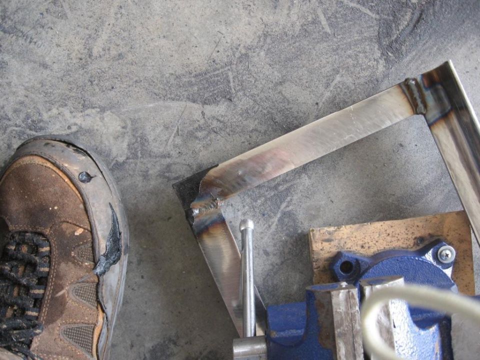 Welding project on metal pieces held in a vise, with a worn work boot in the foreground. Supermotors.net logo visible
