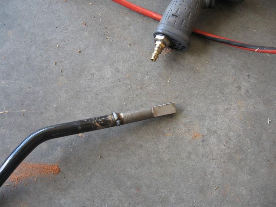 Metal rod with a welded end and a square tip, next to an air-powered tool on a concrete floor
