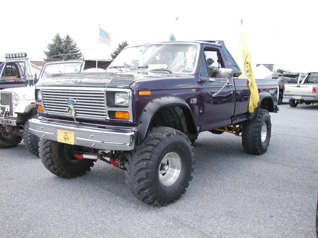 Purple lifted Ford pickup truck with large tires and a yellow flag. Another lifted Jeep is to its left