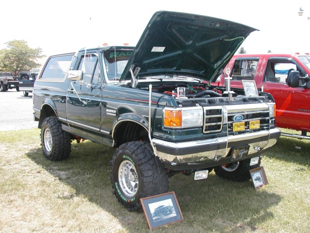 Green Ford Bronco with hood open, lifted suspension, large tires, and chrome grille. A red truck is parked to its right