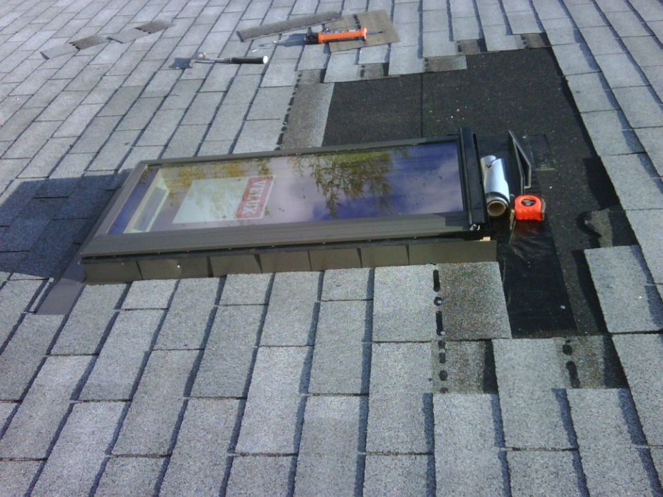 Roof repair in progress showing a skylight installation with shingles, tar paper, tape measure, and hammer