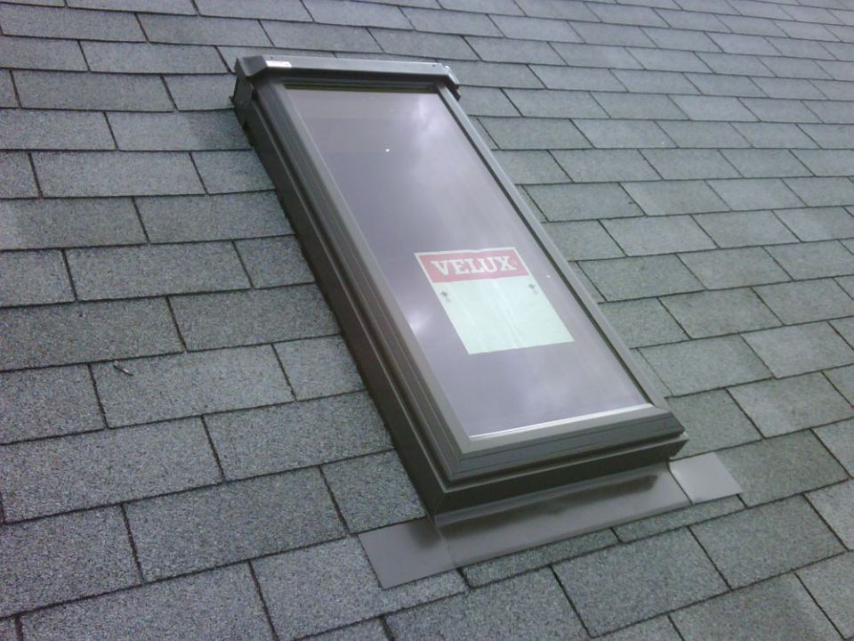 Skylight installed on a gray shingled roof. A VELUX sticker is visible on the glass