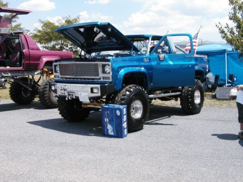 1994 Ford Bronco - A lifted blue pickup truck is displayed with its hood open, alongside another lifted vehicle at an outdoor event.