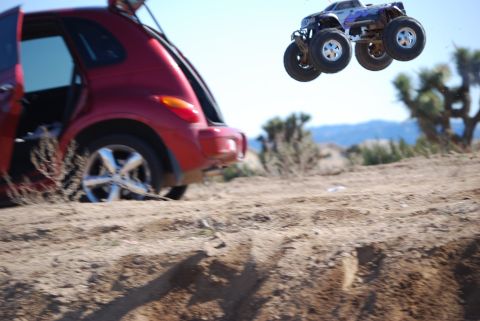 1990 Ford Bronco - RC monster truck performs a jump near a red PT Cruiser in a desert landscape.