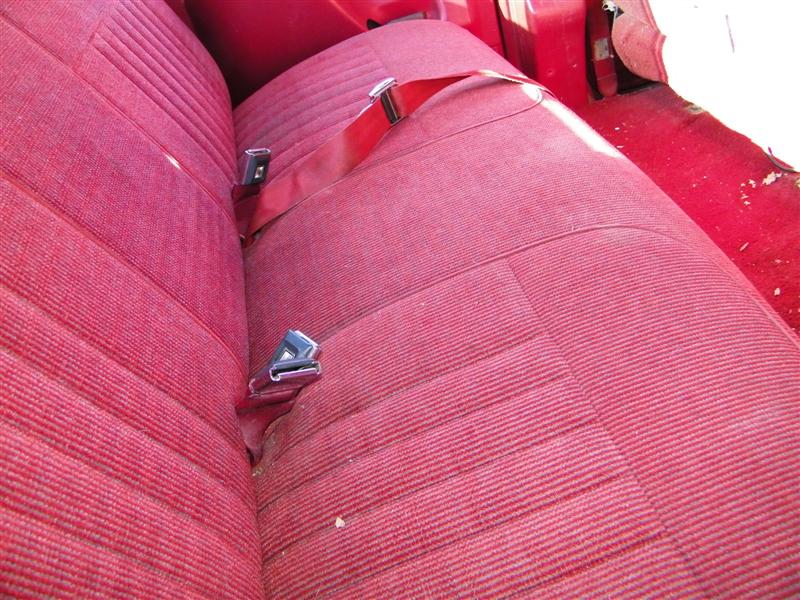 Red fabric bench seat with two red seatbelts and chrome buckles. Visible carpet and part of a white interior panel
