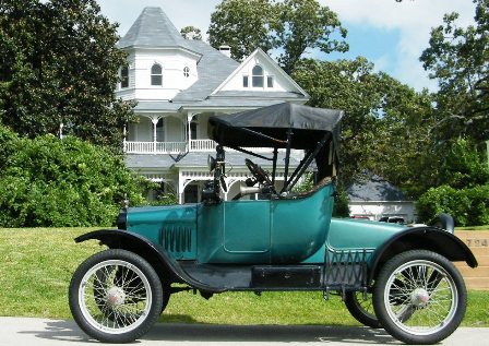 Green Ford Model T roadster in side profile with wire wheels, parked before a white Victorian house