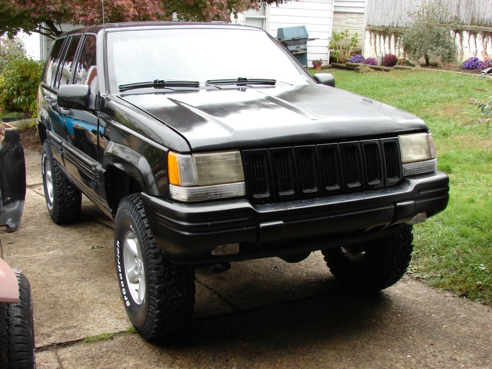 Black Jeep Grand Cherokee parked on a driveway, front quarter view. It has aftermarket tires and a blacked-ou…