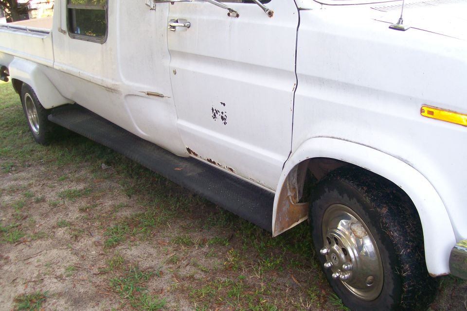 Side view of a white van showing a running board, rust spots, and a portion of the front fender and wheel