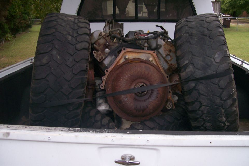 Engine and two large tires secured with straps in the bed of a white pickup truck