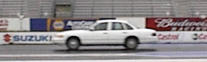 1996 Ford Crown Victoria - A white 1996 Ford Crown Victoria sedan is seen in profile on a race track with various sponsor banners in the background.