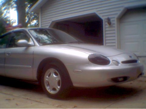 1997 Ford Taurus - A silver sedan is parked in front of a residential garage. The vehicle has a rounded design and tinted headlights.