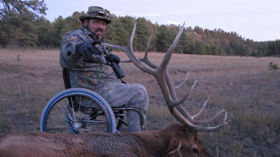 Man in wheelchair points towards camera next to a harvested elk with large antlers