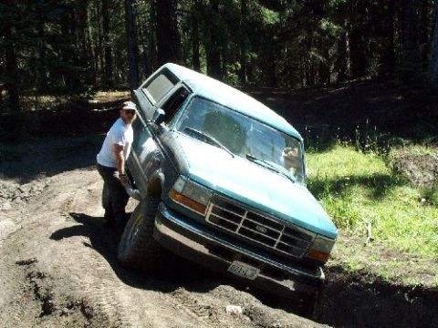 1982 Ford Bronco - A man attempts to free a blue pickup truck with a camper shell that is stuck in a muddy ditch.