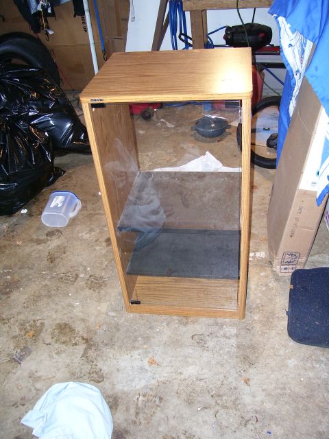 1991 Ford Bronco - A wooden display cabinet with glass doors sits on a concrete floor in a garage, surrounded by various items.