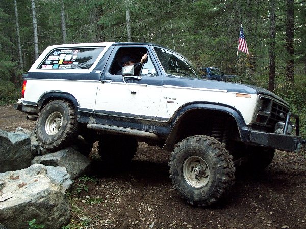 Lifted white and blue Ford Bronco crawling off boulders on a forest trail, driver leaning out