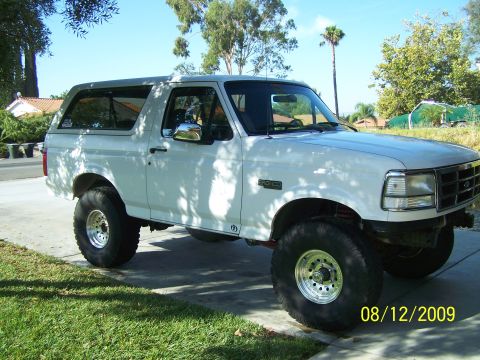 1995 Ford Bronco - White 1995 Ford Bronco with aftermarket wheels and suspension sits parked on a sunny day.