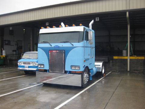 1981 Kenworth W900A - A blue Peterbilt 359 semi-truck is parked next to a smaller blue vehicle in front of a warehouse loading dock.