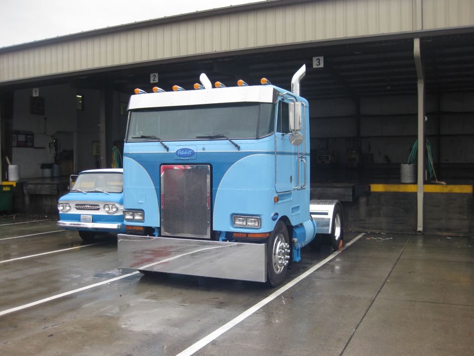 Light blue Peterbilt cabover day-cab tractor with chrome stacks parked outside shop bays