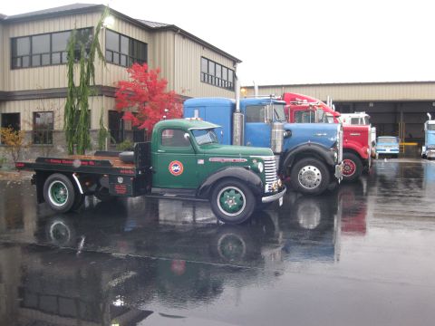 1981 Kenworth W900A - A line of trucks, including a green flatbed, are parked outside a building on a wet day.