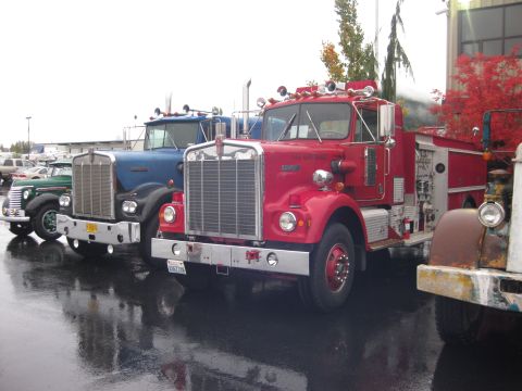 1981 Kenworth W900A - Three vintage trucks, including a red fire truck and a blue semi-truck, are parked in a line on a wet day.
