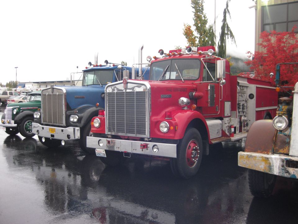 Three vintage trucks parked in a row on a wet surface. The closest is a red fire truck, followed by a blue semi-truck, and a green truck in the background