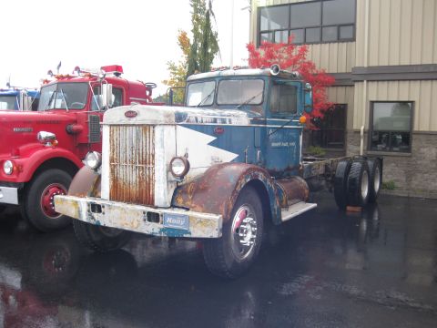 1981 Kenworth W900A - A weathered blue and white truck chassis sits on a wet asphalt lot next to a red truck.
