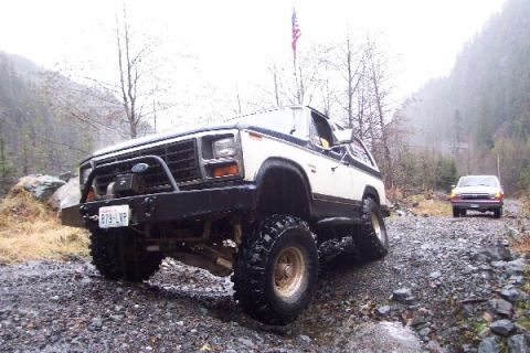 1982 Ford Bronco - 1982 Ford Bronco navigating a rough, wet, gravel road with another vehicle following.