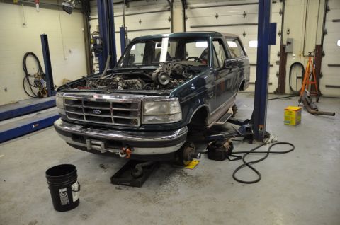 1996 Ford Bronco - A dark green 1996 Ford Bronco is raised on a hydraulic lift in a garage, with its hood open to reveal the engine compartment.