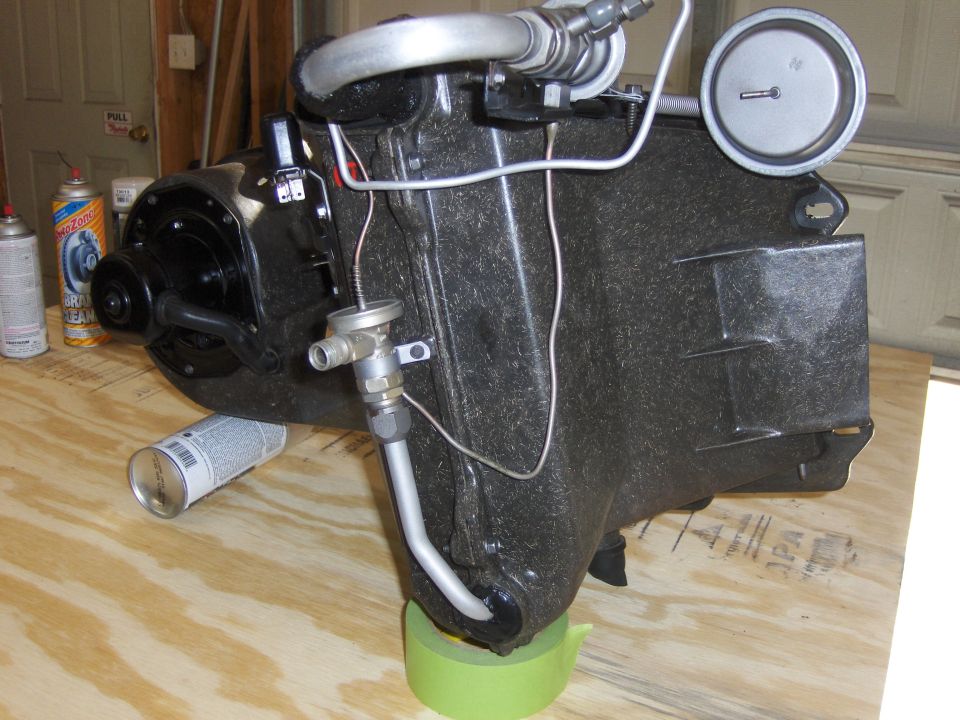 Black textured automotive HVAC evaporator case with metal tubing and a gauge, resting on a wooden surface