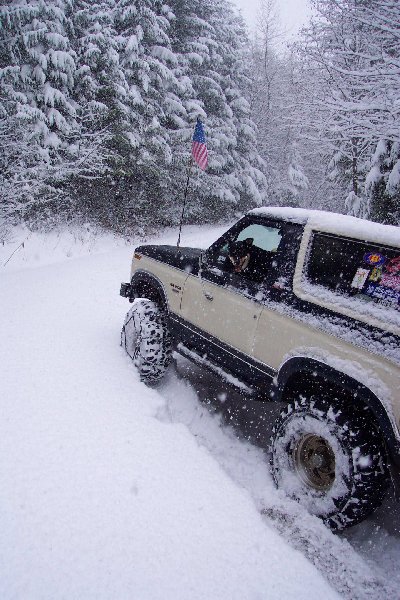 1982 Ford Bronco - A two-tone Ford Bronco navigates a snowy road with snow chains on its tires and an American flag flying from an antenna.