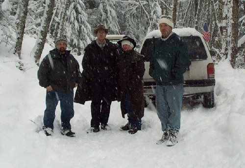 Four people in winter clothing stand in deep snow next to a white SUV with an American flag on its antenna