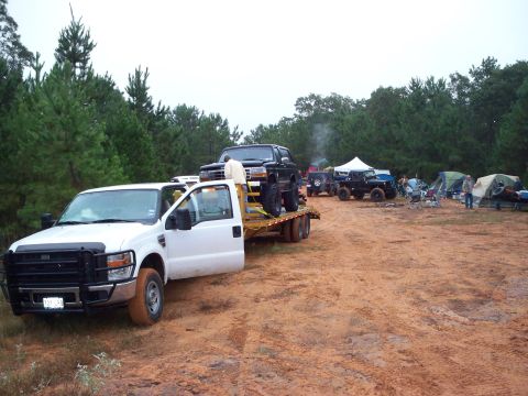 1995 Ford Bronco - A black Ford Bronco is transported on a flatbed truck, with other off-road vehicles and tents visible in the background.