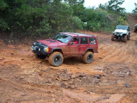 1995 Ford Bronco - A red Jeep Cherokee navigates a muddy off-road trail with other vehicles in the background.