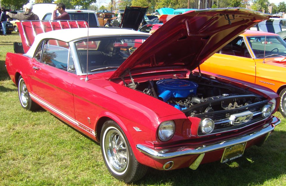 Red 1965 Ford Mustang convertible with white top, hood open showing engine. Another orange car is parked next to it