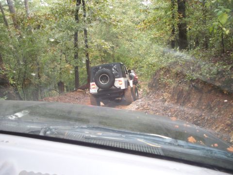 1995 Ford Bronco - Two Jeeps navigate a challenging muddy trail through a dense forest.