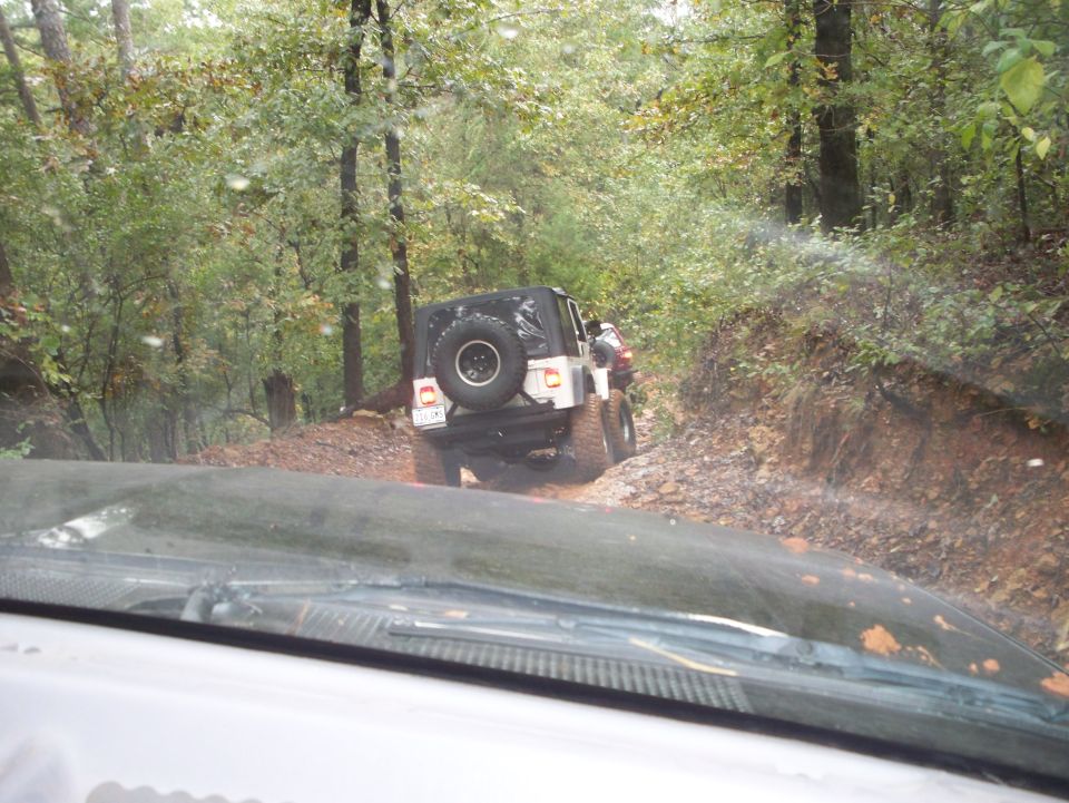 Two Jeeps driving on a muddy, wooded trail. The rear of one Jeep is visible