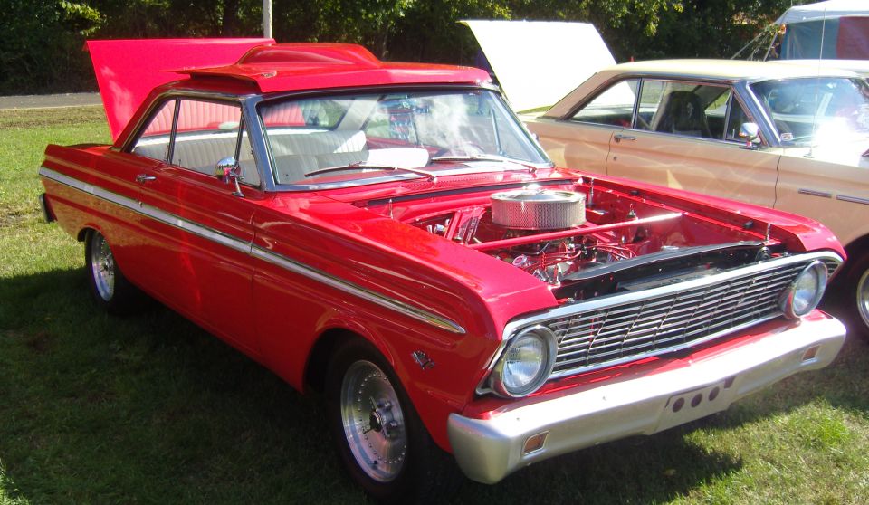Red classic car with hood open, showing engine and chrome grille. Another car is parked behind it