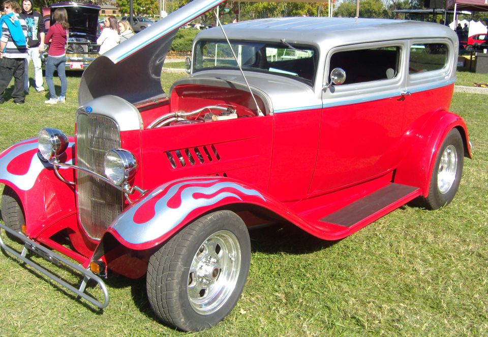 Red and silver hot rod sedan with silver flames on fenders, hood open showing engine, parked on grass