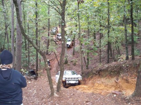 1995 Ford Bronco - Several off-road vehicles traverse a wooded area with a prominent muddy incline.